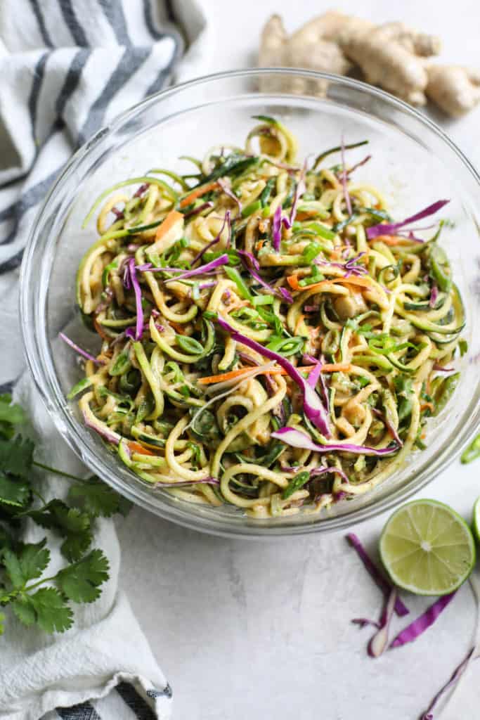 Zucchini noodles with cabbage, carrots, onion in glass bowl surrounded by cilantro, ginger and lime.