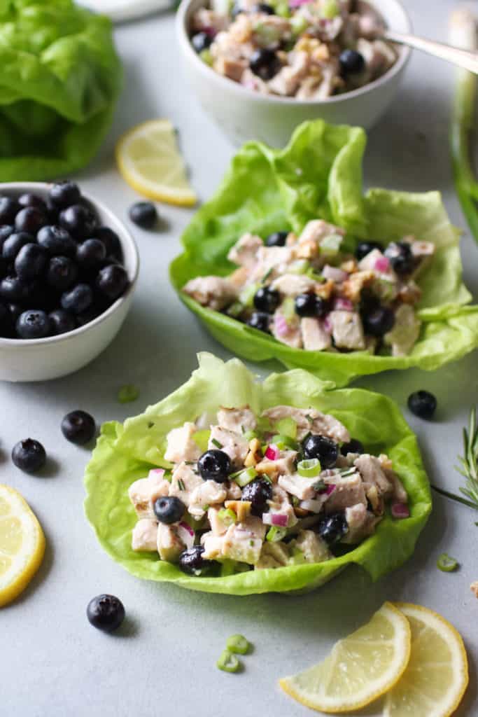Two lettuce cups filled with chicken salad containing blueberries with blueberries on the side and lemon garnish
