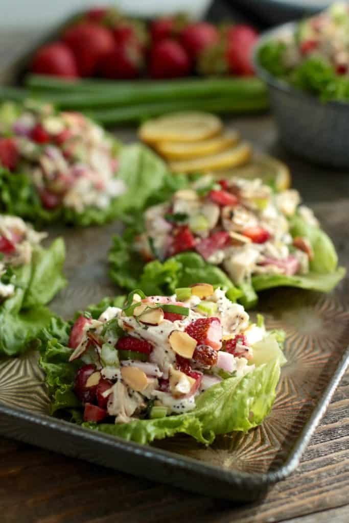 Four lettuce cups with chicken salad, poppy seeds, almonds and strawberries on a pan with onions, strawberries and lemons in the background