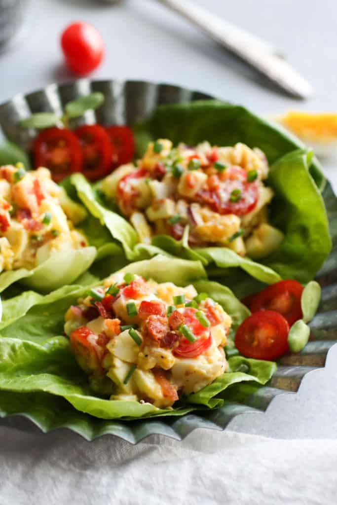 Three lettuce cups filled with egg salad containing tomatoes and bacon on stainless steel tray