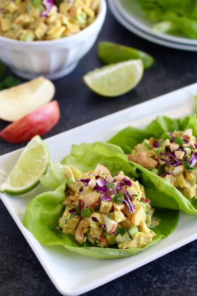 Two lettuce wraps with curry chicken salad on plate topped with apples and cabbage surrounded by apple and lime slices and a bowl of curry chicken salad in the background 