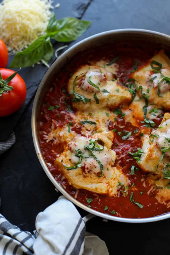 Skillet with tomato sauced and chicken breasts covered in cheese with tomatoes, cheese and basil outside the pan.