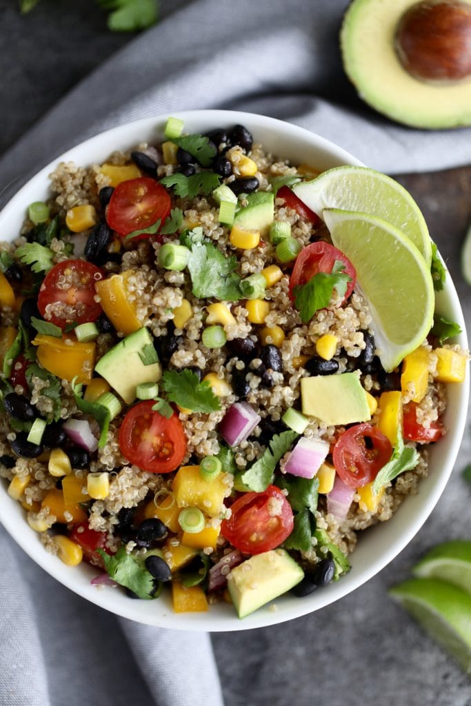 Quinoa salad that includes onion, tomatoes, peppers, avocado and cilantro in a white bowl with lime garnish
