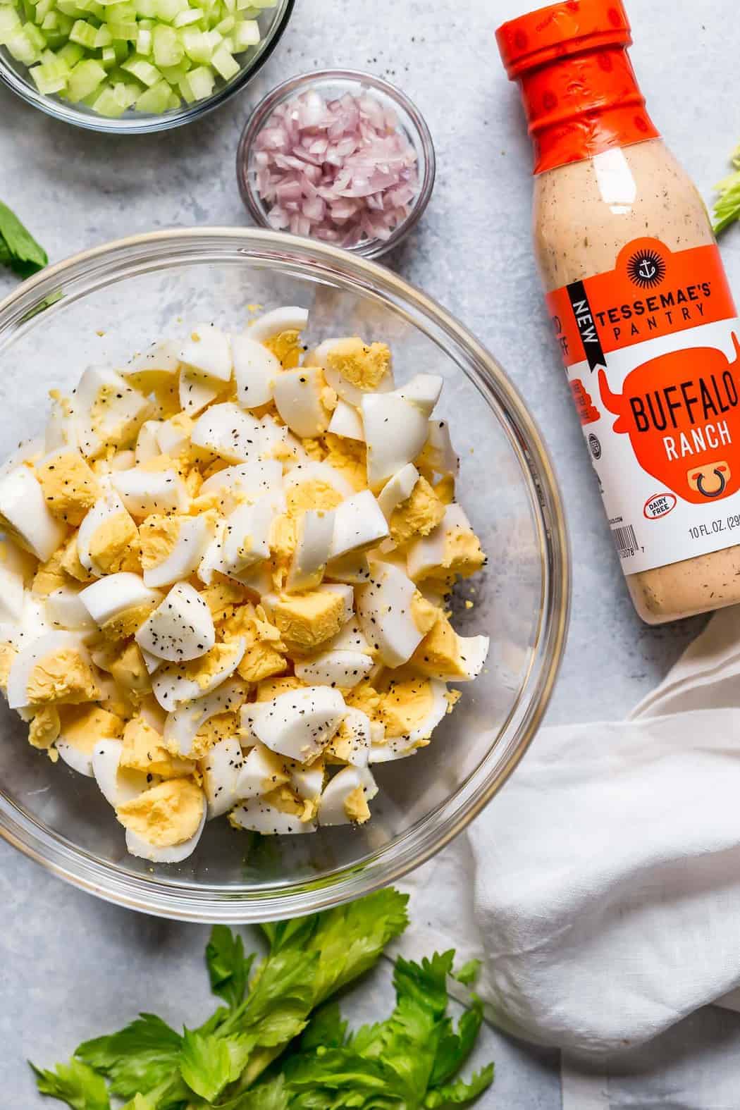 Overhead photo of ingredients for the Buffalo Ranch Egg Salad including a clear bowl of chopped hard-boiled eggs, a clear bowl of minced shallots, a clear bowl of minced celery, a bunch of cilantro and a bottle of Tessemae's Buffalo Ranch Dressing.