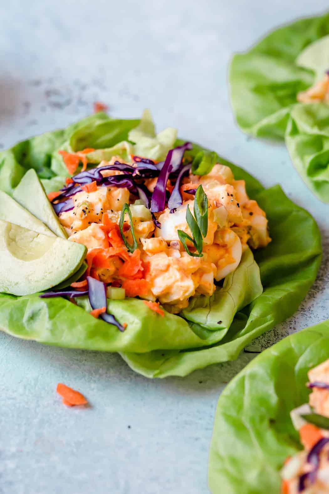 Photo of Buffalo Ranch Egg Salad in a butter lettuce leaf with avocado slice and topped with shredded red cabbage, shredded carrots and green onion slices.