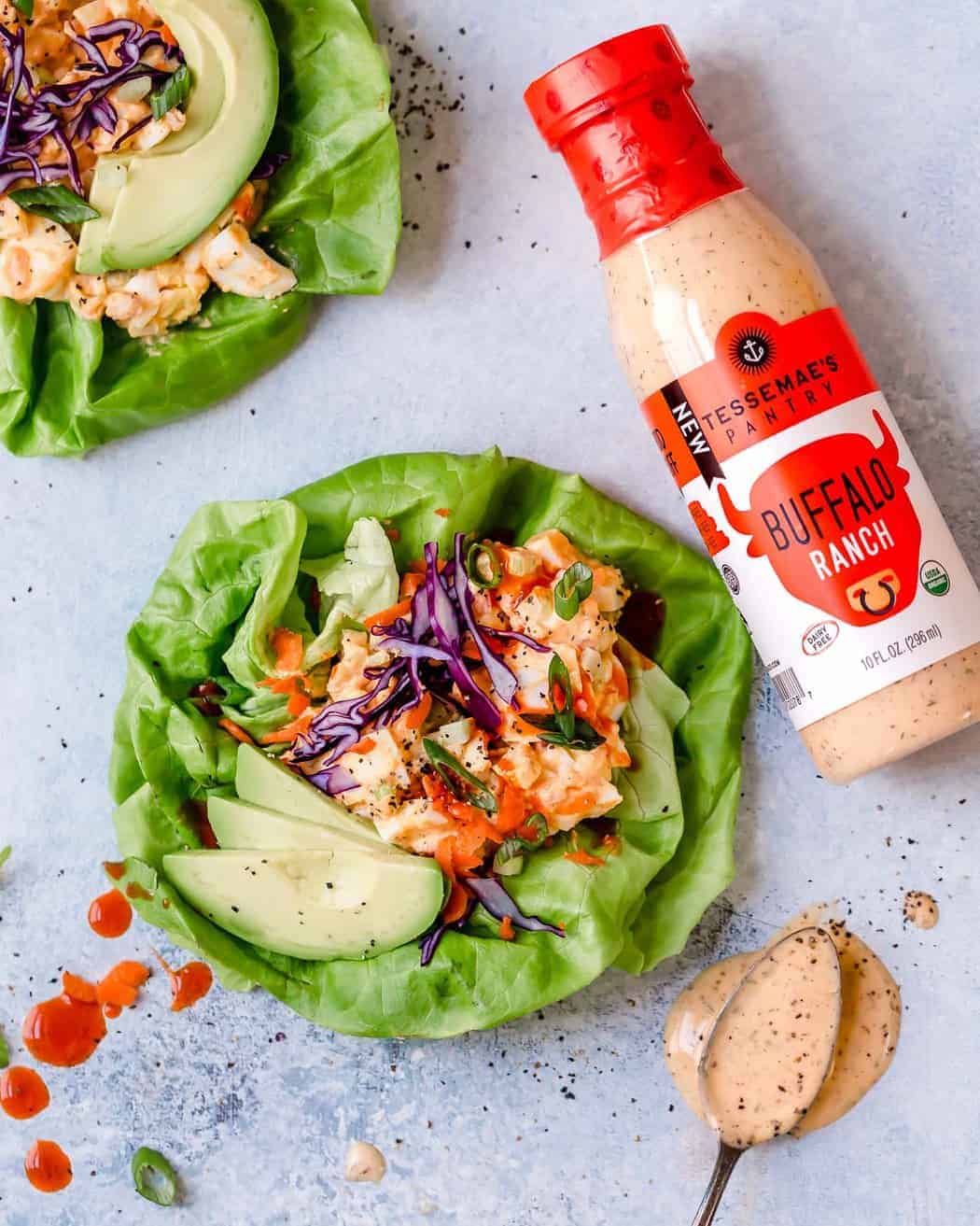 Photo of Buffalo Ranch Egg Salad in a butter lettuce leaf with avocado slice and topped with shredded red cabbage, shredded carrots and green onion slices. Bottle of Tessemae's Buffalo Ranch also in the photo.