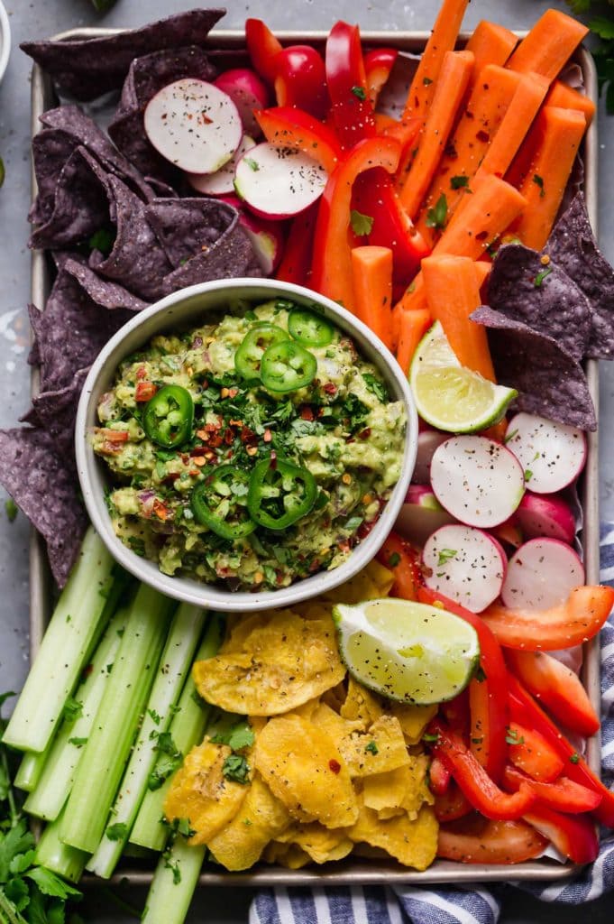 A rainbow of veggies (carrots, celery, red peppers, radishes), plantain and blue corn chips surrounding a bowl of guacamole topped with jalapeños.