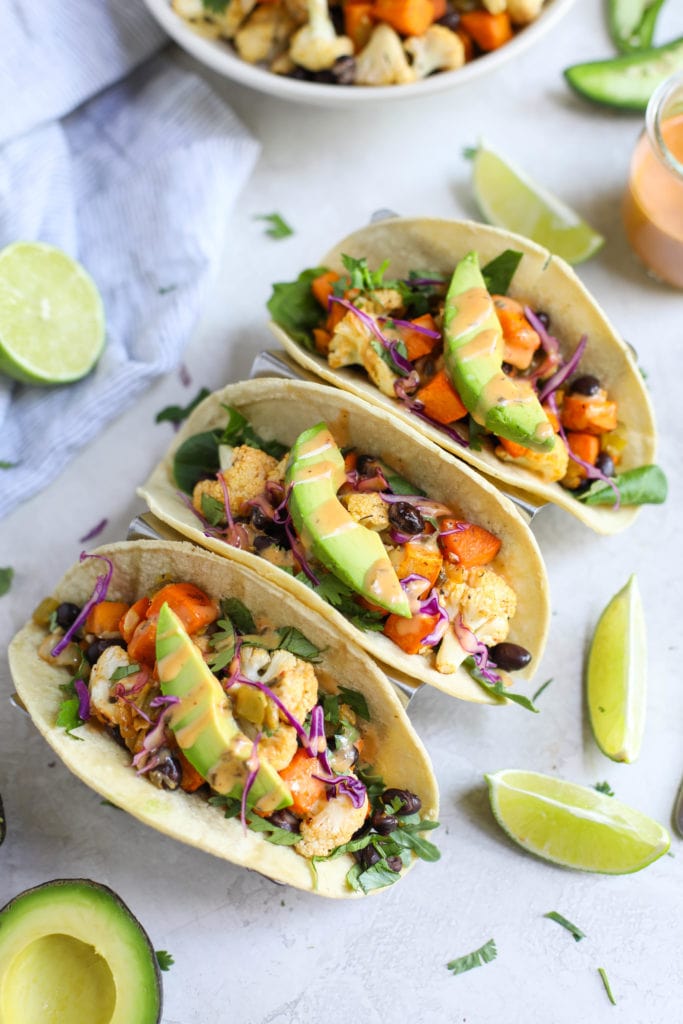 Three soft-shell tacos filled with roasted sweet potatoes and cauliflower drizzled with dressing and surrounded by limes and avocado on white background