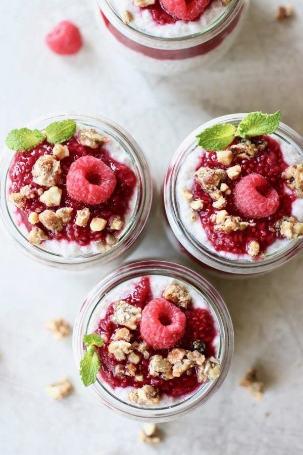 Overhead close-up photo of Raspberry Chia Pudding Parfait divided into 3 small, tulip shaped mason jars each topped with a dollop of raspberry sauce, a fresh raspberry, a few crumbles of granola and a fresh mint leaf.