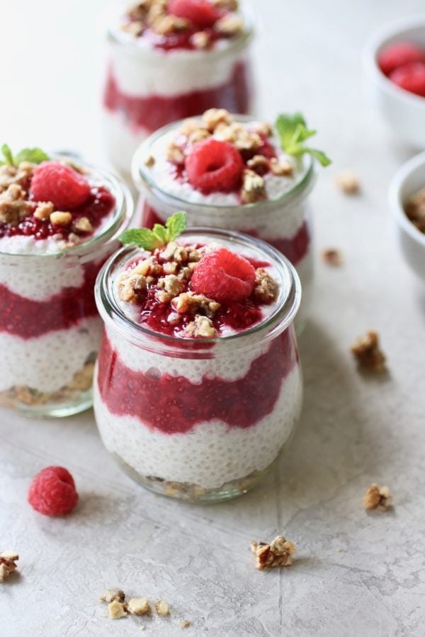 Photo of Raspberry Chia Pudding Parfait dividing in 4 mason jars. Layered with granola at the bottom, chia pudding next, then raspberry sauce, another layer of chia pudding, and a dollop of raspberry sauce on top. Sprinkled with granola and garnished with a mint sprig and fresh raspberry.