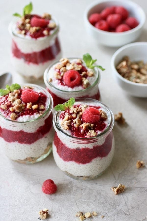 Photo of Raspberry Chia Pudding Parfait divided into 4 small, tulip shaped mason jars. Layered with granola at the bottom, chia pudding next, then raspberry sauce, another layer of chia pudding, and a dollop of raspberry sauce on top. Sprinkled with granola and garnished with a mint sprig and fresh raspberry.