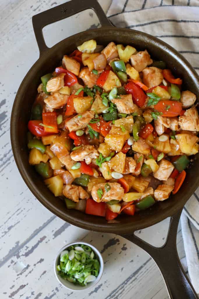 Iron skillet with chicken, peppers, sesame seeds and onions sitting on a striped towel with a side of onions in a small bowl