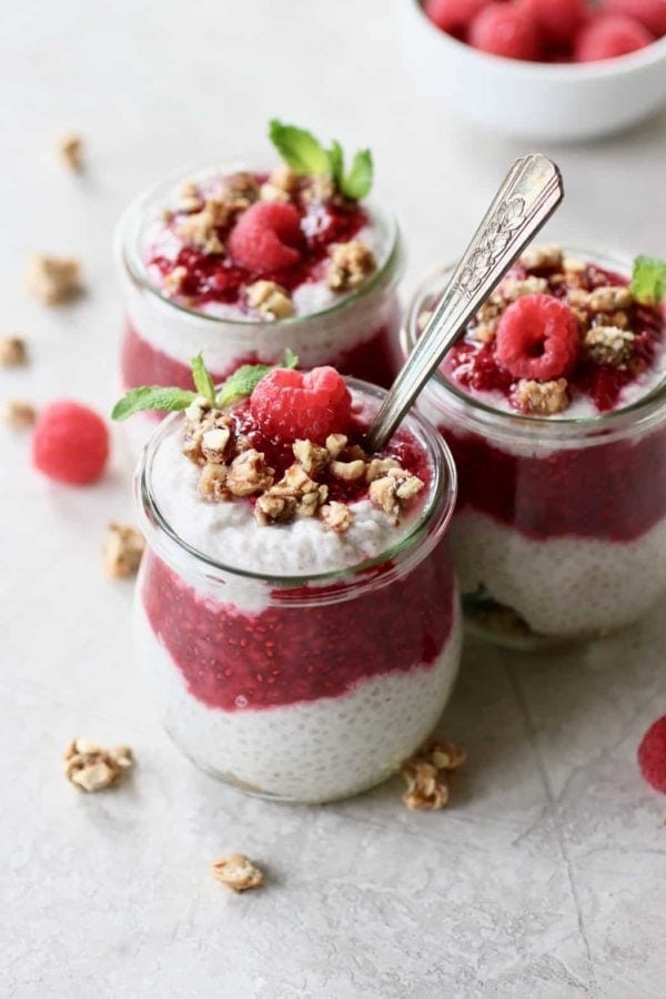 Photo of Raspberry Chia Pudding Parfait divided into 3 small, tulip shaped mason jars. Layered with granola at the bottom, then chia pudding, then raspberry sauce, another layer of chia pudding, and a dollop of raspberry sauce on top. Sprinkled with granola and garnished with a mint sprig and fresh raspberry.