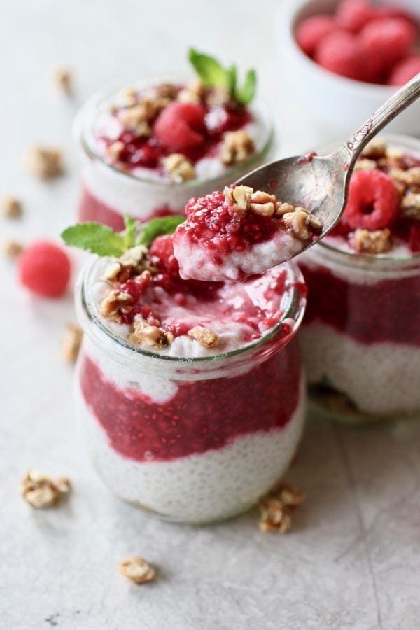 Photo of Raspberry Chia Pudding Parfait divided into 3 small, tulip shaped mason jars. Layered with granola on the bottom, then chia pudding, then raspberry sauce, another layer of chia pudding, and a dollop of raspberry sauce on top. Sprinkled with granola and garnished with a mint sprig and fresh raspberry.