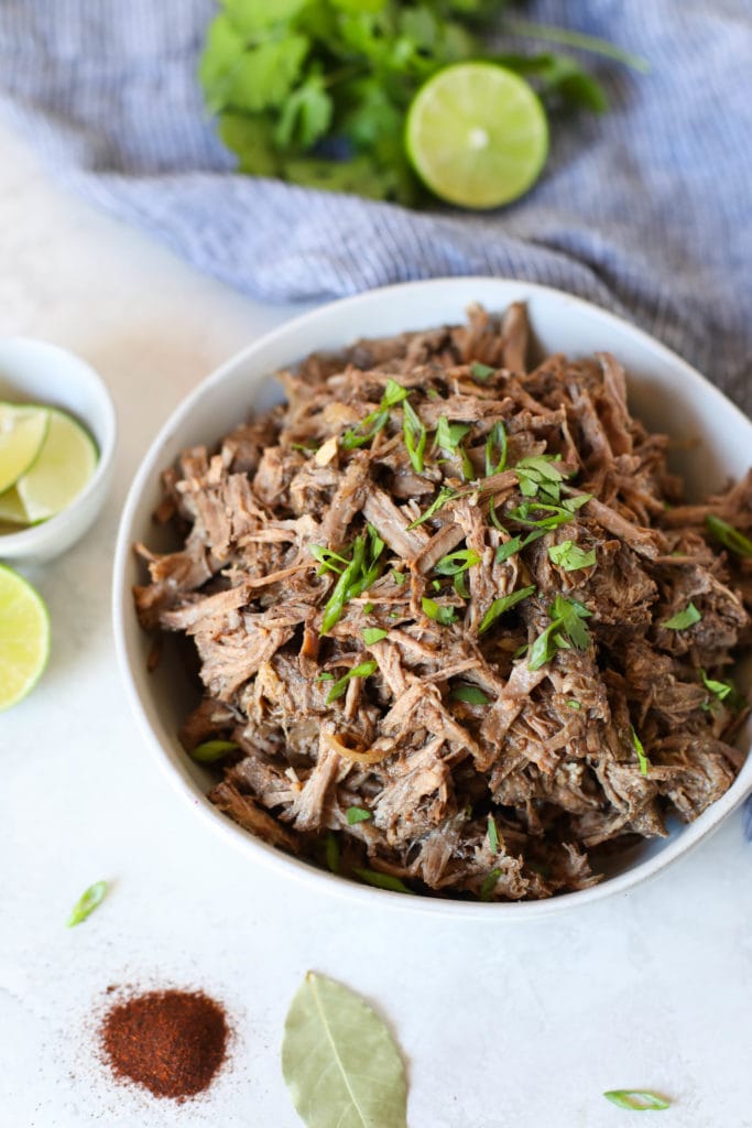 White bowl filled with shredded barbacoa beef with green onions on top and garnished with lime and cilantro 