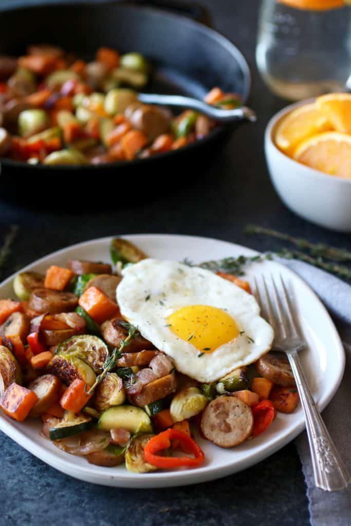 Plate of sliced chicken sausage, zucchini, sweet potato, red peppers, Brussels sprouts topped with an egg with a fork on plate and an iron skillet with more hash in the background as well as a side of orange slices