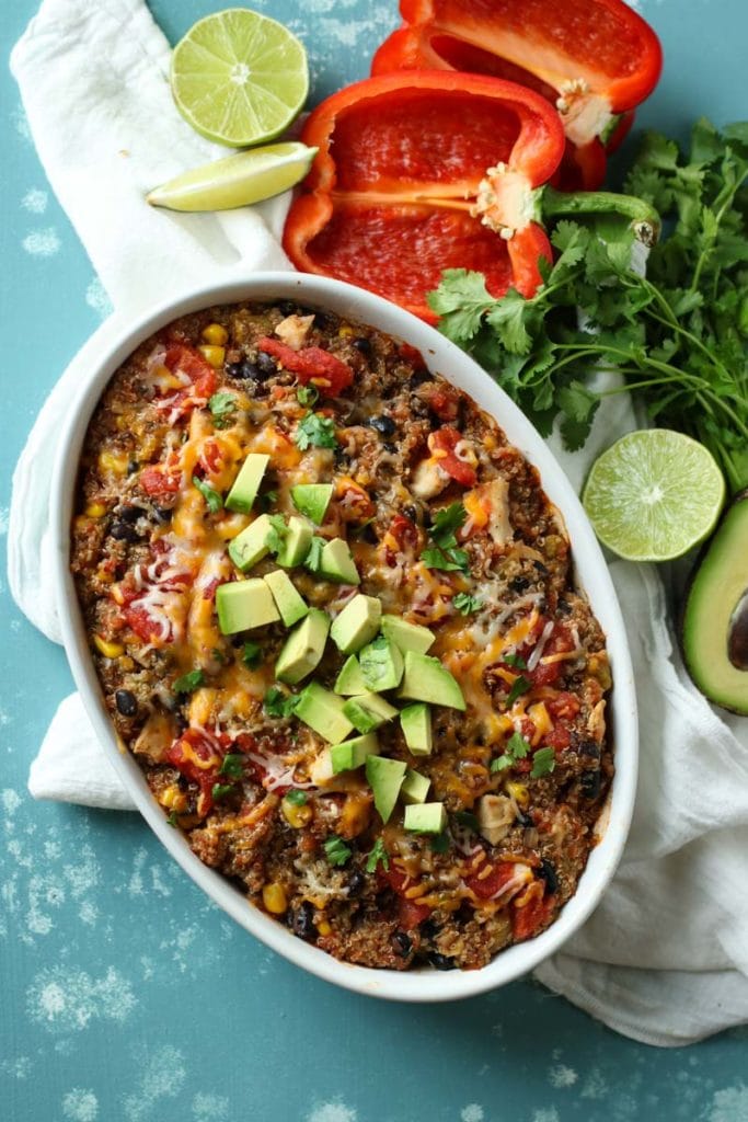 Oval white baking dish with quinoa bake topped with avocado and surrounded by limes, red peppers, limes and cilantro.