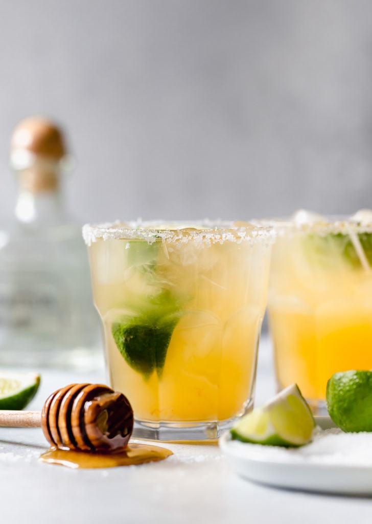 Two margaritas in salted tumblers with a honey-dipped wand laying flat and lime garnishes and a tequila bottle in the background