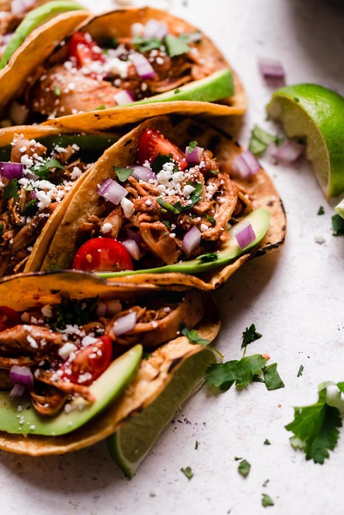 Folded tacos in corn tortillas with avocado, tomatoes, onions and chicken with limes and cilantro surrounding them.