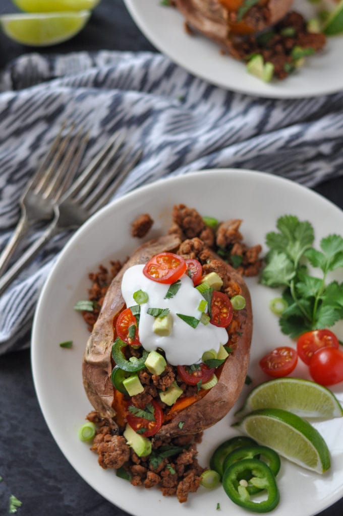 Sweet potato stuffed with taco meat and topped with jalapeño peppers, tomatoes, avocado and sour cream on a white plate garnished with more peppers, limes, tomatoes and cilantro. Forks, napkin and another plate of food in background.