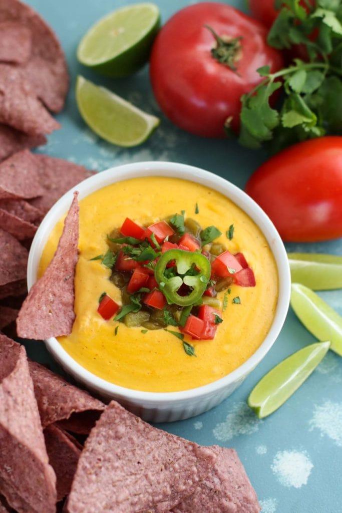 A white bowl filled with nacho cheese, a blue chip and pepper garnish surrounded more with blue corn chips, tomatoes, and lime garnish