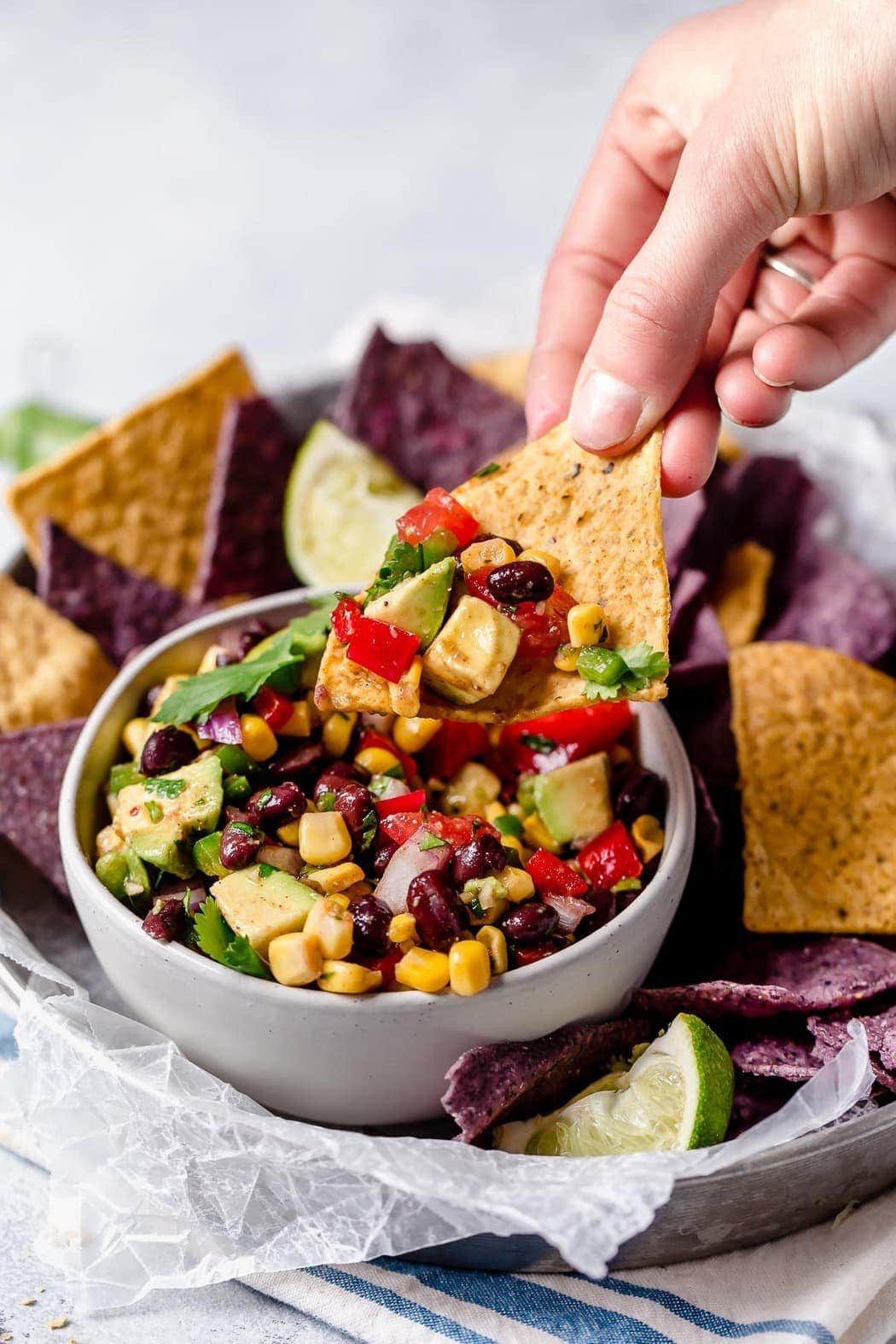 Black Bean & Corn Salsa with Avocado in a white bowl surrounded by blue and white corn tortilla chips. Showing a hand picking up a chip with a scoop of salsa on it.