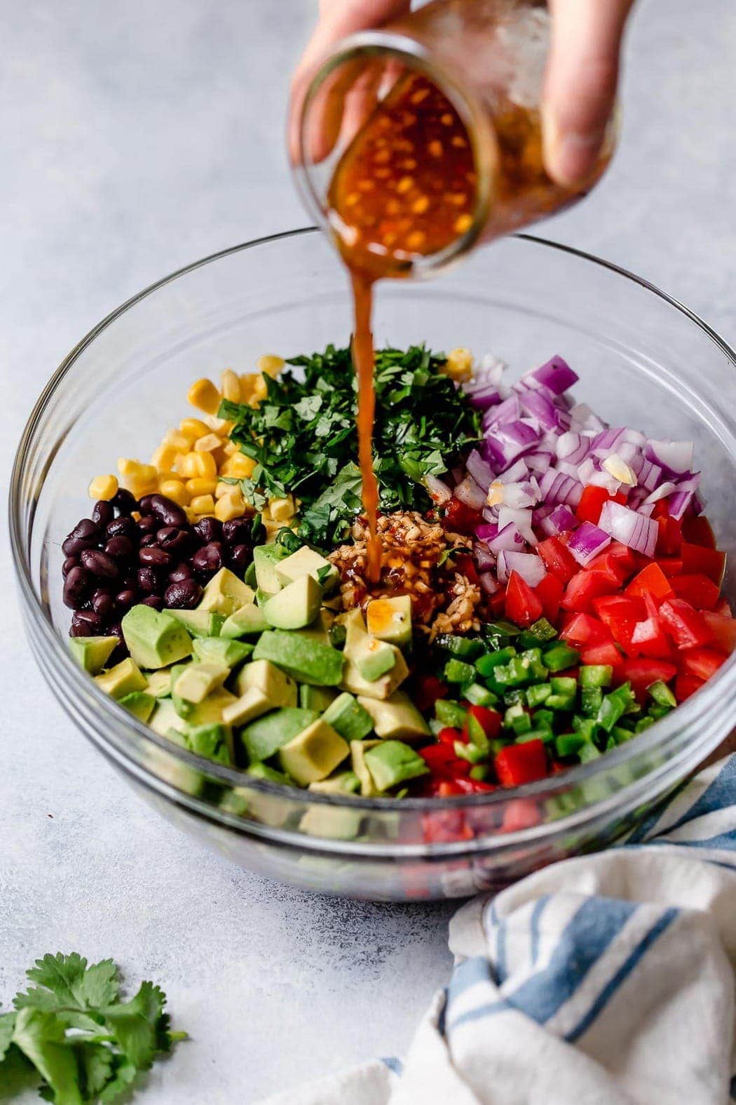 Black Bean & Corn Salsa with Avocado in a clear bowl before it has been mixed showing the chili lime vinaigrette being poured onto the salsa ingredients. 