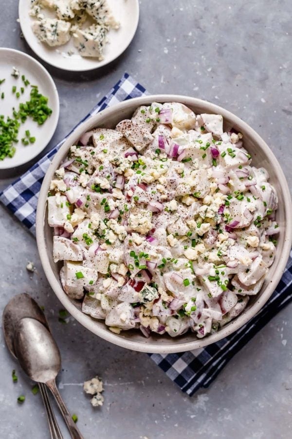 Big bowl filled with potato salad on blue and white checkered napkin with spoon by it and two white plates with chives and blue cheese by it. 