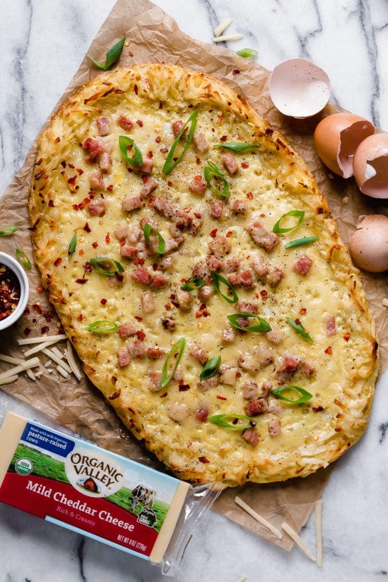 Overhead photo of Breakfast Pizza with Hash Brown Crust topped with green onion slices and crushed red pepper. Also included is a block of Organic Valley Cheddar Cheese and brown egg shells.