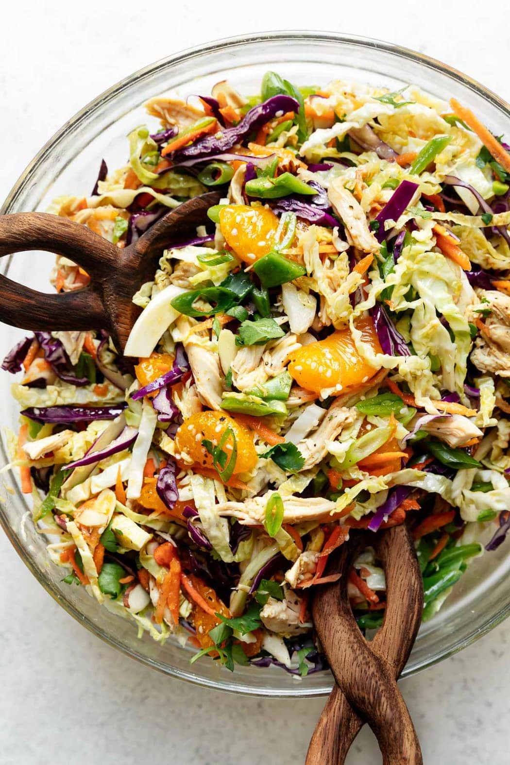 A large serving bowl filled with mandarin orange chicken salad with two wooden handled salad spoons in the salad.