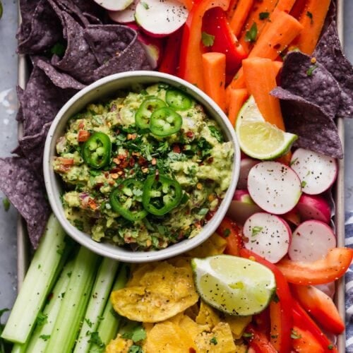Overhead view of a bowl of fresh guacamole surrounded by colorful cut vegetables and blue corn tortilla chips.