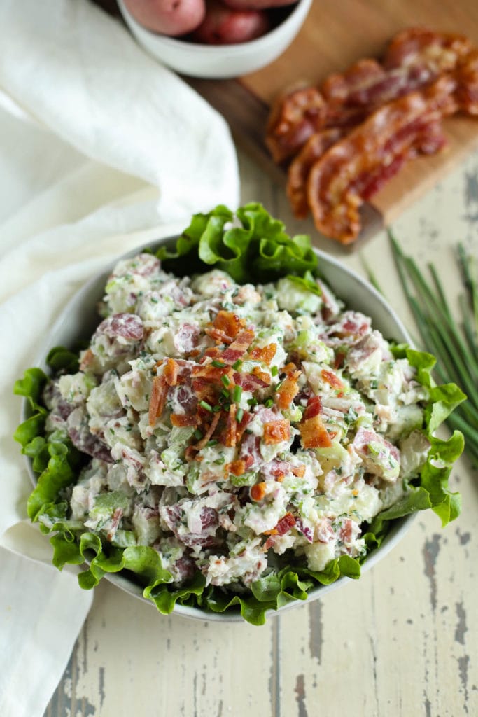 Lettuce-lined bowl filled with potato salad with bacon crumbled on top with potatoes and bacon in background on weathered white surface.