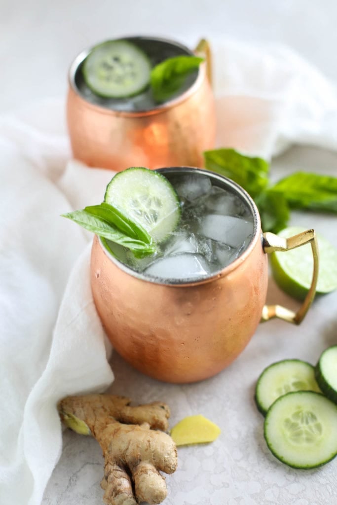 Two drinks in copper mugs filled with cucumber, mint and ice. Lime and cucumber slices resting around the,