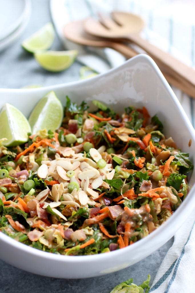 White bowl filled with chopped salad with brussels sprouts, carrots, bacon, almonds and golden raisins with lime wedges on side