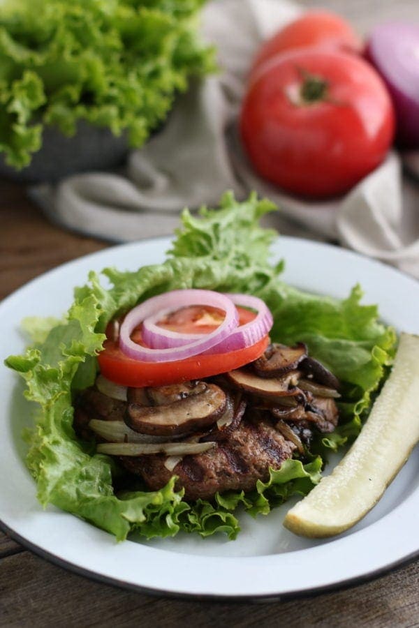 Grilled burger topped with mushroom, tomato and onions and wrapped in lettuce leaf on a white plate with a pickle spear on the side.