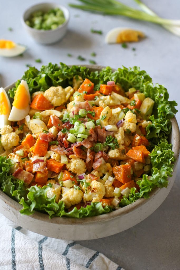 Bowl of lettuce-lined bowl filled with sweet potatoes, cauliflower salad with bacon and hard-boiled egg on side