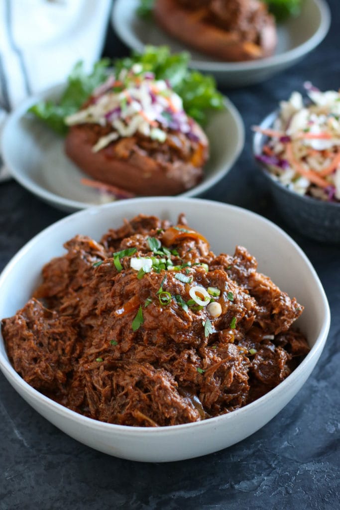 White bowl of bbq shredded beef garnished with onions. With plated bbq beef stuffed sweet potato in background