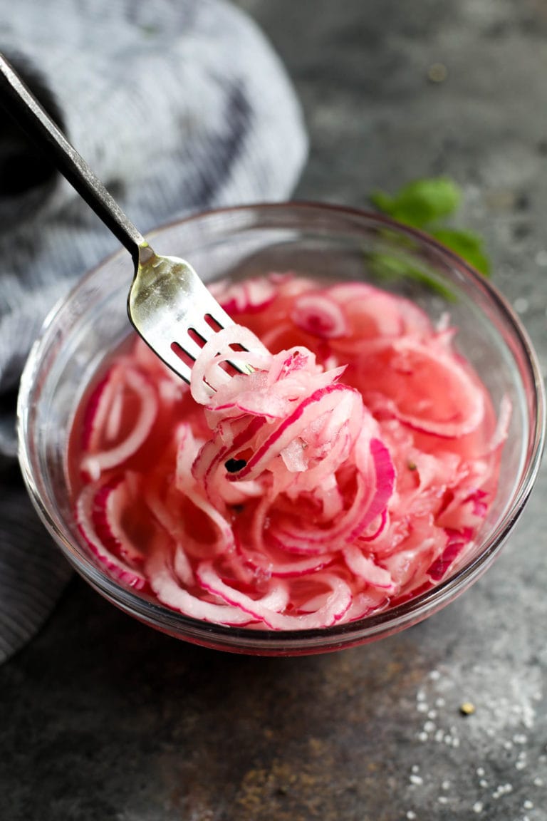 A fork lifting sliced pickled red onions from a mixing bowl filled with pickled red onions.