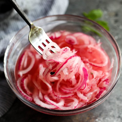 A fork lifting sliced pickled red onions from a mixing bowl filled with pickled red onions.