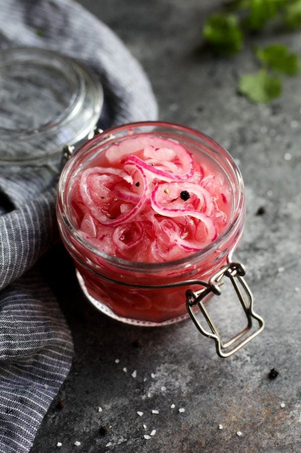 Quick pickled red onions in a small glass jar topped with a few peppercorns.