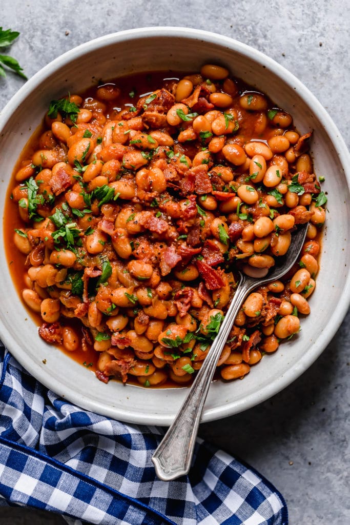 Bowl of baked beans with bacon chunks and herb garnish with a spoon dipped in it and blue-checker napkin around it