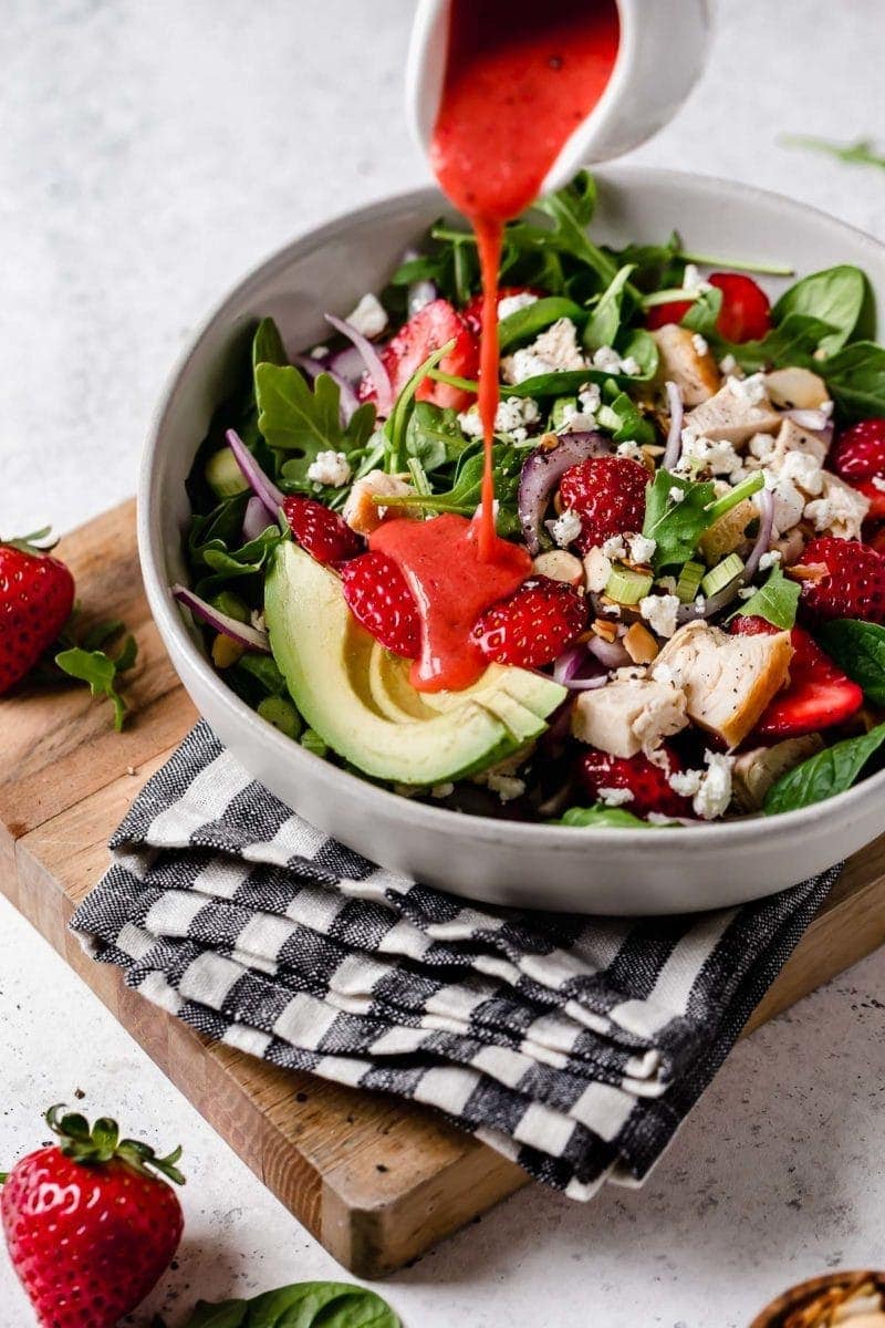 Red strawberry vinaigrette being poured over strawberry spinach salad with avocado and chicken pieces, in stone bowl