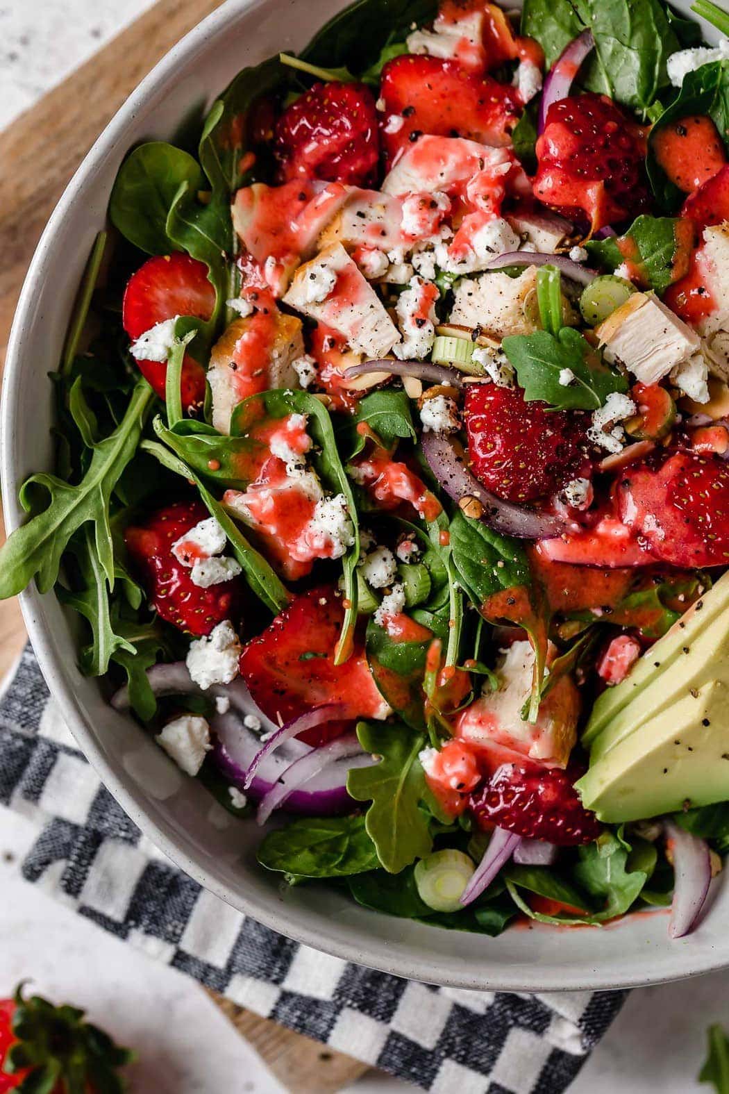 Gorgeous closeup overhead shot of Strawberry Spinach Salad with Chicken in a white bowl. Ingredients include greens, sliced strawberries, chunks of chicken, goat cheese crumbles, avocado slices and red onion slices. 