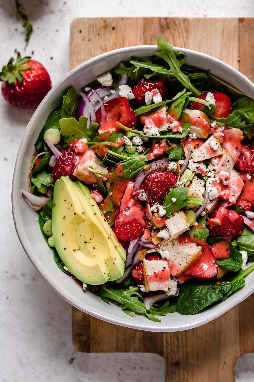 Gorgeous overhead shot of Strawberry Spinach Salad with Chicken in a white bowl. Ingredients include greens, sliced strawberries, chunks of chicken, goat cheese crumbles, avocado slices and red onion slices. 