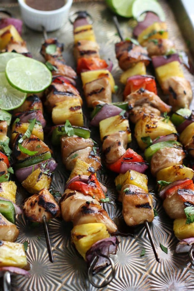 Six chicken kebabs threaded with chicken, pineapple, peppers and onions on a sheet pan with a small dish of dressing toward the back