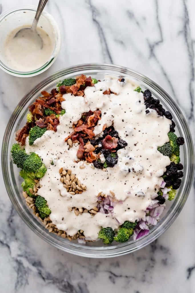 Creamy yogurt dressing poured over top of all ingredients for creamy broccoli salad in mixing bowl
