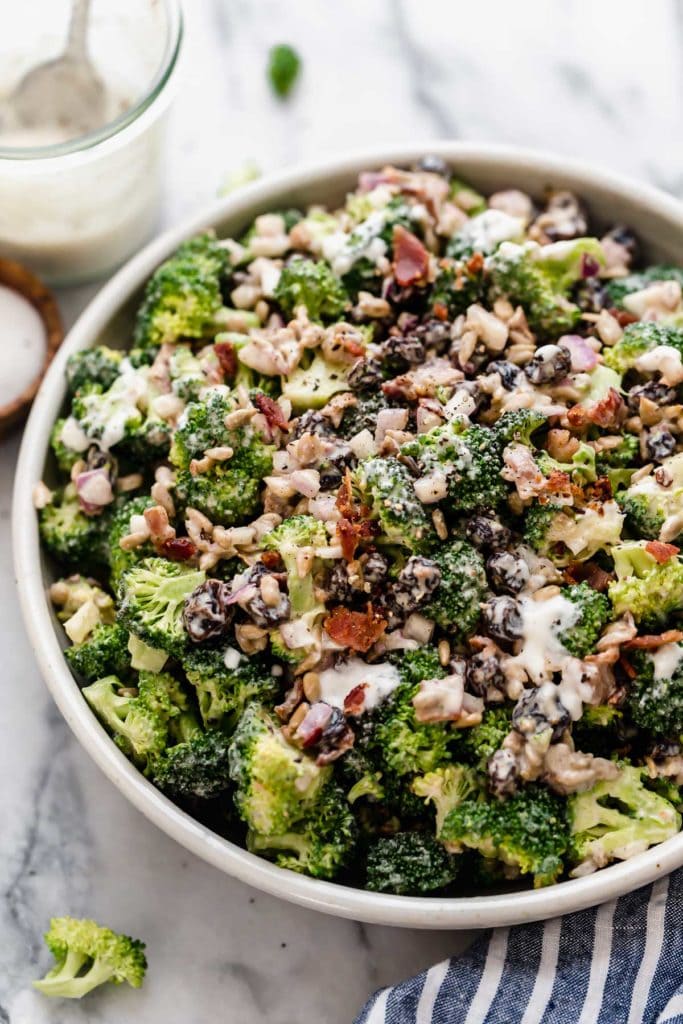 Bowl filled with broccoli salad with sunflower seeds, craisins and bacon with dressing drizzled over the tip. Glass container with a spoon partially filled with dressing by bowl