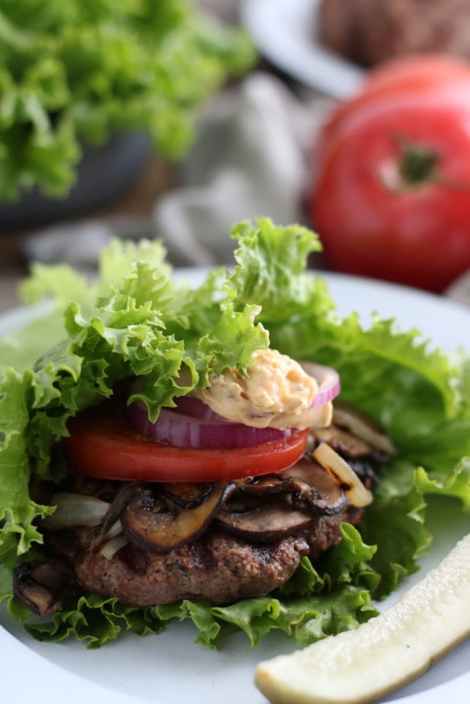 Grilled burger topped with mushrooms, onions, tomatoes onion and mayo wrapped in a lettuce leaf on a plate. Tomato and lettuce in the background