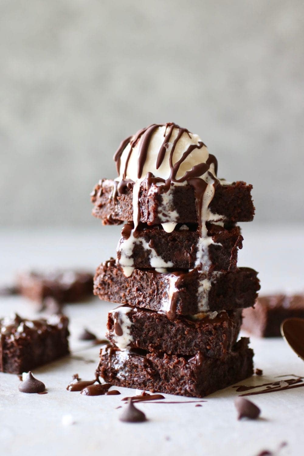 Stack of five fudgy brownies topped with a melting scoop of vanilla ice cream. Other brownies and chocolate chips surrounding them.