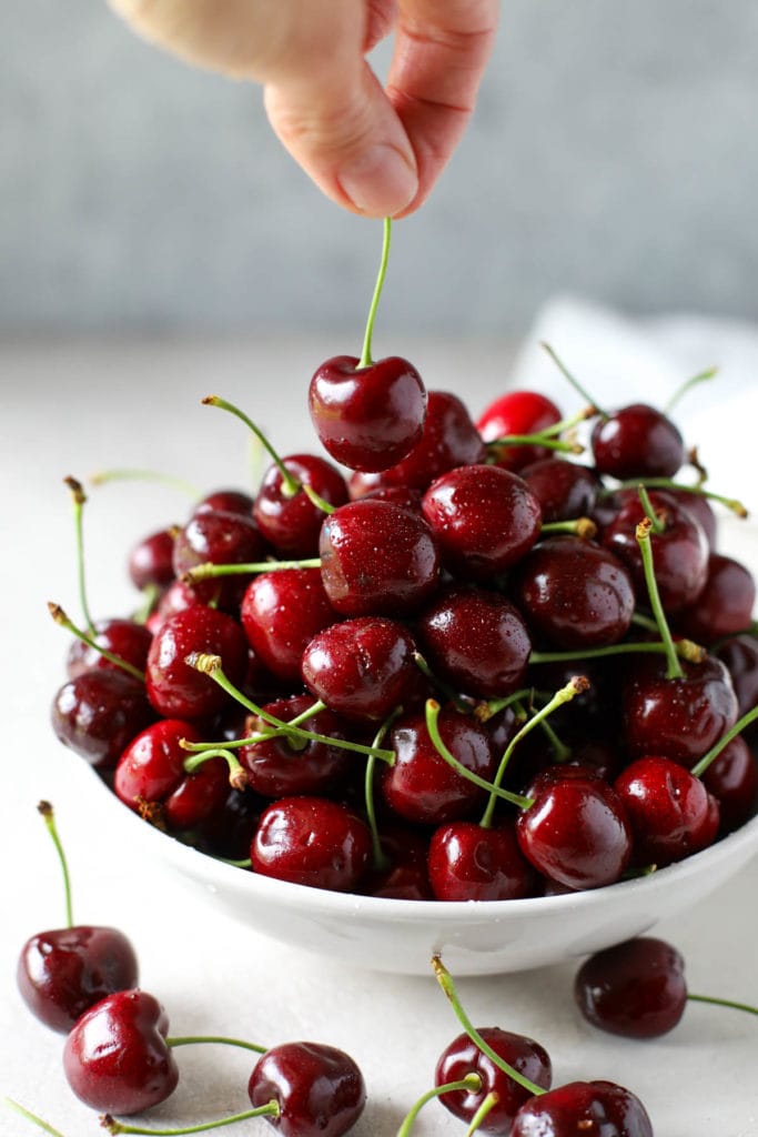 Bowl of cherries in a white bowl with two fingers pulling one cherry out.
